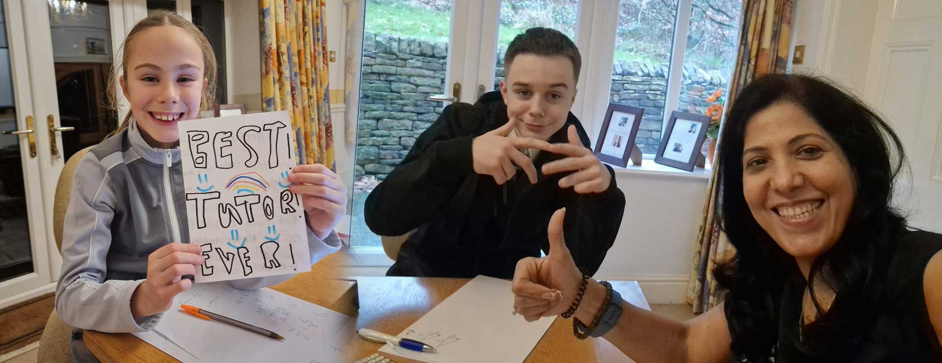 Neelu with two of her pupils at the kitchen table, one holding up a hand-drawn 'Best tutor ever!' sign